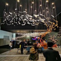 A stunning large custom lighting hotel chandelier dominates a modern space. This big chandelier features an array of suspended glass feathers, transitioning from white to golden hues, creating a sense of movement like migrating birds. Thin wires and twinkling lights add a celestial effect. Below, additional custom lighting elements include floating disc-like structures. People gather beneath, admiring the chandelier that transforms the hotel lobby into an immersive, artistic wonderland.