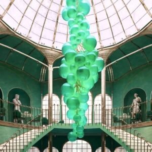 A stunning custom lighting installation featuring a large hotel chandelier composed of numerous mint green glass spheres. This big chandelier cascades from the center of a domed glass ceiling, creating a whimsical, bubble-like formation. The custom lighting piece contrasts beautifully with the rich emerald green walls and ornate architecture of the atrium. Below, a circular bar area is visible, while the upper levels showcase statues in alcoves. The chandelier's ethereal glow and unique design make it a captivating centerpiece in this luxurious, multi-level space, blending modern artistry with classical elements.