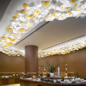 A stunning custom lighting installation featuring a large chandelier that borders the ceiling of an upscale dining area. This hotel chandelier comprises numerous illuminated glass elements shaped like abstract petals or leaves, in shades of white and warm yellow. The big chandelier creates a flowing, organic pattern around a central rectangular recess in the ceiling. Below, a circular buffet area is visible, adorned with various dishes and a plant centerpiece. The warm, textured walls complement the soft glow of the chandelier, creating an inviting and luxurious atmosphere for diners.