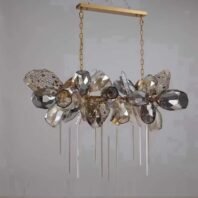 A striking linear chandelier showcasing innovative custom lighting design. The fixture features a horizontal gold-toned metal bar suspended by two chains. From this bar hangs a cluster of diverse geometric crystal-like elements in various shapes, sizes, and textures. The palette includes clear, smoky gray, and champagne-colored pieces. Some elements are faceted and smooth, while others have intricate filigree or honeycomb patterns. The arrangement is asymmetrical and organic, creating a sense of controlled chaos. Beneath the main cluster, multiple slender crystal rods hang vertically at varying lengths, adding a delicate, icicle-like quality to the design. This luxurious custom lighting piece would serve as a captivating focal point over a dining table or in a grand entryway, perfect for those seeking a blend of contemporary geometry and organic forms in their lighting design.