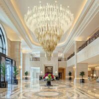 A breathtaking large chandelier dominates the luxurious lobby of an upscale hotel. This custom lighting masterpiece features multiple tiers of elongated crystal-like elements, creating a cascading effect. The top layers appear in clear or white tones, gradually transitioning to golden hues towards the bottom, forming an elegant ombre design. The chandelier's massive scale perfectly complements the grand, two-story lobby with its ornate ceiling, marble floors, and classic architectural details. Ambient lighting around the ceiling's perimeter enhances the chandelier's glow. The opulent space is further adorned with potted plants, arched windows, and a central floral arrangement. This stunning centerpiece exemplifies high-end hotel design, seamlessly blending contemporary lighting artistry with traditional luxury aesthetics.