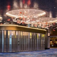 An awe-inspiring large hotel chandelier showcasing innovative custom lighting design. This big chandelier spans across multiple circular ceiling panels, creating a floating garden effect. Hundreds of glass elements resembling flowers and leaves in white and coral hues cascade from the illuminated discs, appearing to drift through the air. The intricate arrangement extends the length of a curved hallway, complementing the modern architecture with its flowing glass doors. Soft ambient lighting from the chandelier bathes the space in a warm glow, exemplifying how custom lighting can transform a hotel's interior into a mesmerizing work of art.