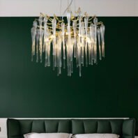 A luxurious crystal chandelier with icicle-like pendants and gold accents hanging above a dark green tufted headboard. The chandelier features numerous elongated crystal drops suspended from gold-toned branches, creating a waterfall effect. The fixture is illuminated, casting a warm glow through the crystal elements. The chandelier is set against a deep green wall, creating a striking contrast between the sparkling crystals and the rich background color. Only the top of the green tufted headboard is visible at the bottom of the image, suggesting a bedroom or upscale living space setting. The combination of the chandelier's delicate design and the bold green color scheme creates an atmosphere of sophistication and opulence.