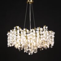 A luxurious oval-shaped chandelier featuring a multitude of white and clear crystal discs suspended from gold-toned branches. The chandelier is illuminated, creating a warm glow that radiates through the translucent discs. It's hung from a circular gold ceiling mount by thin wires. The crystal elements vary in texture, with some appearing smooth and others having a dimpled surface, creating a dynamic visual effect. The overall design resembles a cluster of illuminated bubbles or snowflakes, blending modern aesthetics with classic opulence. The chandelier is photographed against a dark background, emphasizing its intricate details and the interplay of light through its structure.