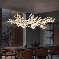 A contemporary dining room featuring a striking horizontal bubble cluster chandelier. The chandelier consists of numerous white frosted glass orbs and golden accents, creating an organic, cloud-like formation above a long dining table. The fixture's warm light contrasts beautifully with the room's dark, moody ambiance. The dining area includes a sleek black table surrounded by mid-century modern style chairs with brown leather seats and backs. The room's design incorporates various textures: a textured stone wall to the left, wood paneling, and smooth dark walls. A metal and wood shelving unit is visible in the background. The space exudes a sophisticated, modern aesthetic, balancing warmth and coolness through its lighting and material choices.