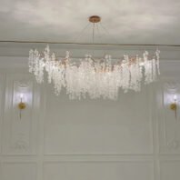 Luxurious rectangular crystal chandelier with golden accents suspended in a classical-style room. The chandelier features numerous textured crystal disc pendants cascading from golden branches, creating an organic, cloud-like form. It's suspended from a round gold ceiling plate by thin wires. The room has ornate white wall panels with decorative moldings and two matching wall sconces with crystal accents flanking the chandelier. A golden picture rail runs along the top of the walls. The ceiling appears to have a subtle textured or painted finish. The chandelier's warm glow illuminates the space, highlighting the interplay between the modern fixture and the traditional architectural elements.