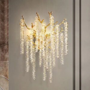 A luxurious wall-mounted chandelier featuring golden branches and cascading crystal strands. The fixture's structure consists of organically shaped gold-toned metal branches that extend outward, mimicking tree limbs. Hanging from these branches are multiple strands of small, clear crystals arranged in long, vertical formations resembling icicles or delicate chains. The crystals catch and refract light, creating a warm, shimmering effect against the light-colored wall background. The chandelier is illuminated, casting a soft, golden glow that highlights both the metallic and crystal elements. The piece is mounted on what appears to be a neutral-toned wall, with part of a decorative wall panel or mirror frame visible to the right. The chandelier's design blends natural, organic forms with glamorous materials, resulting in a striking and elegant lighting fixture suitable for upscale interior spaces.