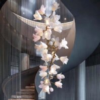 Breathtaking branch chandelier adorns a contemporary stairwell, featuring delicate white and pink flower-shaped lights. Golden branches twist elegantly, supporting numerous illuminated blooms. The chandelier is set against a backdrop of shimmering string curtains and curved architectural elements. This stunning branch chandelier cascades down the stairwell, creating a harmonious blend of nature-inspired design and modern luxury. The soft glow of the floral lights contrasts beautifully with the sleek, grey interior, adding warmth and sophistication to the space.