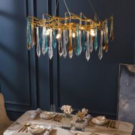 A sophisticated dining room featuring a stunning chandelier with colorful glass icicle-like droplets. The chandelier hangs above a marble-topped dining table set for a meal. The fixture consists of clear, blue, and amber glass pieces suspended from golden, branch-like metal structures. Illuminated bulbs within the chandelier cast a warm glow, highlighting the unique textures of the glass elements. The room has dark blue walls with decorative molding, creating a rich backdrop for the chandelier. The dining table is elegantly set with white plates, gold cutlery, and wine glasses. Decorative coral-like sculptures in clear cases serve as centerpieces. In the background, a portion of an abstract artwork is visible, adding to the room's modern yet classic ambiance. The overall scene combines luxury and artistry, with the chandelier serving as a striking focal point in this sophisticated dining setting.