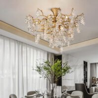 A luxurious dining room featuring a stunning chandelier that resembles a golden tree branch with cascading crystal clusters. The chandelier hangs above a partially visible dining table set with plates and glasses. Below, a large vase with green branches adds a natural touch to the space. The room has white walls, sheer curtains, and hints of marble, creating a bright and sophisticated atmosphere. The chandelier serves as a focal point, blending organic forms with opulent materials in a modern, yet nature-inspired design.