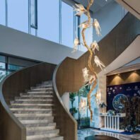 A contemporary lobby featuring a striking vertical chandelier shaped like a golden branch with illuminated glass birds. The chandelier extends from floor to ceiling, complementing a curved marble staircase. Large windows allow natural light to flood the space. To the right, a decorative blue panel with floral motifs adds a touch of traditional elegance. The space blends modern architecture with nature-inspired design elements, creating a unique and inviting entrance area.