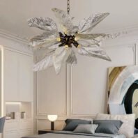 A sophisticated bedroom featuring a striking chandelier with large, translucent glass leaves arranged in a flower-like pattern. The chandelier hangs from a white ceiling with ornate molding. Below, a bed with gray pillows is visible. White walls with decorative paneling create a classic backdrop. A modern abstract artwork hangs on the wall, balancing traditional and contemporary elements. The room's design showcases a blend of natural-inspired lighting and refined interior decor.