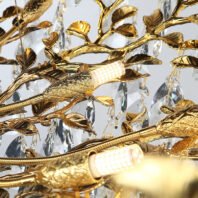 Detailed view of a gold-plated chandelier with intricate leaf designs and crystal droplets. Illuminated LED lights are visible within gold bird-shaped fixtures. The branches intertwine, creating a nature-inspired motif with sparkling crystal elements.