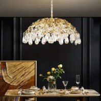 A sophisticated dining space featuring a stunning gold leaf and crystal dome chandelier as the focal point. The chandelier hangs above a modern wooden dining table set with elegant dinnerware and gold-toned cutlery. The room has a dramatic black paneled wall backdrop, creating a striking contrast with the illuminated chandelier. A wood art piece with an abstract, curved design adorns the wall, adding warmth and texture. A vase of fresh yellow roses sits on the table, complementing the gold tones of the chandelier and place settings. The overall ambiance is one of luxury and contemporary style, blending organic shapes with metallic accents.