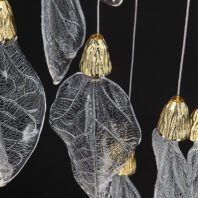 Close-up of a sophisticated branch chandelier featuring delicate glass leaves suspended from thin wires. Each leaf-shaped element is intricately textured to mimic real foliage, with visible veins and a translucent quality that catches the light beautifully. The leaves are crowned with ornate gold bell-shaped caps, adding a luxurious touch to the nature-inspired design. This branch chandelier combines organic forms with refined materials, creating a stunning interplay of light and shadow. The contrast between the clear, textured glass and the polished gold accents exemplifies a perfect balance of natural elegance and modern luxury in lighting design.