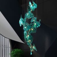 Elegant branch chandelier featuring a vertical arrangement of illuminated teal glass leaves, suspended in a modern stairwell. The chandelier's design mimics a flowing vine with glowing foliage, each leaf-shaped element lit from within. Set against a dark wall, the teal glass creates a striking contrast. The chandelier's organic form complements the sleek, geometric lines of the staircase, blending nature-inspired artistry with contemporary architecture. A potted plant below echoes the chandelier's botanical theme.