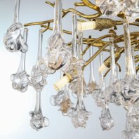 A detailed view of an exquisite chandelier combining gold-plated metalwork with intricate glass elements. The upper part of the image shows a network of curved, gold-toned metal arms forming the chandelier's structure. Hanging from these arms are numerous clear glass pendants, each with a unique, organic shape reminiscent of abstract flowers or marine life. The glass elements feature long, slender stems that transition into more elaborate, bulbous forms at their ends. These pendants have a hand-blown appearance with swirling patterns and textures that catch and refract light beautifully. Interspersed among the glass pendants are gold-plated accents with a textured, pineapple-like design. Several small LED bulbs are visible, emitting a warm glow that illuminates the crystal elements. These lights are housed in gold-toned fixtures that blend seamlessly with the overall design. The chandelier's composition creates a stunning interplay between the warm gold tones of the metalwork and the clear, sparkling glass pendants. This piece combines classical elegance with modern, artistic glasswork, resulting in a lighting fixture that serves as both a functional light source and a captivating sculptural element. Such a chandelier would be a luxurious focal point in high-end interior spaces, perfect for dining rooms, grand entryways, or upscale living areas.