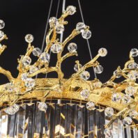 A close-up view of an opulent chandelier featuring intricate gold-plated branches adorned with crystal elements. The image showcases a complex network of golden branches that twist and curve organically, reminiscent of tree limbs or coral formations. These branches extend from a central, textured gold band with an ornate, filigree-like design. Attached to the golden branches are numerous faceted crystal orbs of varying sizes, resembling sparkling dewdrops or blossoms. The crystals are precisely cut, allowing them to catch and refract light brilliantly. Below the gold band, a series of long, rectangular crystal prisms hang vertically, creating a curtain-like effect. These prisms add an additional layer of light refraction and sparkle to the overall design. Thin silver wires can be seen suspending the chandelier, contrasting with the warm gold tones. The chandelier is photographed against a dark background, which emphasizes the luminous quality of the gold and crystal elements. This chandelier beautifully merges nature-inspired design with luxurious materials, resulting in a piece that is both a functional light fixture and a stunning work of art, perfect for creating a dramatic focal point in high-end interior spaces.