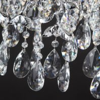 A close-up view of an exquisite crystal chandelier against a dark background, showcasing the intricate details of its design. The image features multiple teardrop-shaped crystal pendants of varying sizes cascading downward. These crystal teardrops are precisely faceted, brilliantly refracting light and creating prismatic effects. Interspersed among the larger teardrops are smaller, round crystal beads connected by thin wires. A notable feature is the presence of silver-toned metal elements shaped like leaves or vines, adding an organic, nature-inspired touch to the design. These metallic leaves contrast beautifully with the transparent crystals. The chandelier components are arranged in a layered, organic pattern, giving the impression of a crystalline waterfall or an elaborate ice formation adorned with silver foliage. Light hitting the various crystal elements produces a dazzling display of sparkles and rainbow-like reflections, which stand out dramatically against the dark background. This detailed view emphasizes the chandelier's luxurious craftsmanship, its ability to create a mesmerizing interplay of light and crystal, and the harmonious blend of geometric crystal shapes with naturalistic metallic accents.