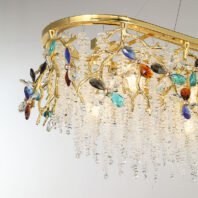 A close-up view of a luxurious oval-shaped chandelier featuring a harmonious blend of gold, clear crystals, and multi-colored gemstone-like accents. The fixture is framed by a polished gold oval rim, from which intricate golden branches extend downward. These branches are adorned with vibrant crystal flowers in various colors including blue, turquoise, amber, and clear, providing striking pops of color against the predominantly gold and clear crystal design. The most captivating feature is the abundant cascade of clear crystal strands hanging from the gold frame, creating a shimmering curtain effect reminiscent of a waterfall or icicles. These crystal strands are arranged in varying lengths, adding depth and dimension to the piece. The clear crystals catch and refract light, producing a dazzling sparkle. Thin wires can be seen suspending the chandelier, adding to its floating, ethereal appearance. This chandelier masterfully combines the warmth of gold with the cool brilliance of crystals and the vibrancy of colored gemstones, resulting in a piece that is both a functional light source and an exquisite work of art, capable of making a dramatic statement in any sophisticated interior space.