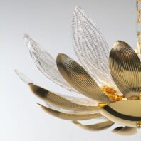 A close-up view of an exquisite branch chandelier featuring a flower-like design with alternating gold and crystal petals. The chandelier's structure resembles a blooming flower, with elongated petals radiating outward from a central golden core. The gold petals have a smooth, ribbed surface with a warm metallic sheen, while the crystal petals are clear and intricately textured with a pattern reminiscent of delicate feathers or leaf veins. The crystal elements catch and refract light, creating a sparkling effect against the light background. The petals are arranged in a layered, overlapping pattern, giving the chandelier a sense of depth and dimensionality. At the center, a golden orb forms the flower's core, from which the petals seem to unfurl. The image captures the chandelier's meticulous craftsmanship, showcasing the seamless blend of opaque gold and transparent crystal elements. A glimpse of the chandelier's chain is visible in the upper right corner, hinting at its suspended installation. This branch chandelier masterfully combines organic, floral-inspired forms with luxurious materials, resulting in a stunning piece that bridges the gap between nature and high-end interior design.