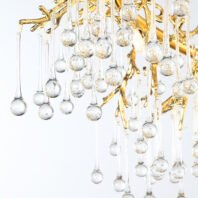 A close-up view of an exquisite branch chandelier featuring golden metalwork and an array of crystal pendants. The chandelier's structure is partially visible, showcasing intricately designed gold-toned branches that resemble tree limbs. Suspended from these golden branches are numerous clear crystal pendants of varying lengths, each consisting of a slender stem ending in a spherical drop. These crystal elements mimic raindrops or dewdrops, creating a cascading effect. Some of the crystal spheres contain delicate gold flecks or bubbles, adding a touch of whimsy and luxury. The image is captured against a light background, emphasizing the transparency of the crystal drops and the warm glow of the gold branches. This branch chandelier masterfully combines organic, nature-inspired forms with opulent materials, resulting in a stunning piece that bridges the gap between natural beauty and high-end interior design.