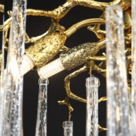 Close-up of a luxurious branch chandelier showcasing intricate gold-plated metalwork. The chandelier features elaborately textured golden branches with lifelike bark patterns and twigs. Delicate crystal pendants, shaped like elongated icicles, hang from the golden structure, creating a stunning contrast. The image highlights the exquisite craftsmanship of the branch chandelier, capturing the interplay between the warm, reflective gold surfaces and the sparkling, transparent crystal elements. Small light fixtures are integrated into the design, visible as golden cylinders with textured patterns. This branch chandelier combines nature-inspired artistry with opulent materials, perfect for adding a touch of elegance and drama to high-end interiors.