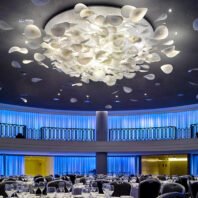 A stunning large chandelier dominates a grand hotel ballroom, showcasing innovative custom lighting design. This big chandelier resembles a cloud of illuminated white petals, creating a mesmerizing focal point against a dark ceiling. The hotel chandelier's organic shape contrasts beautifully with the room's modern blue-lit perimeter and sleek balcony. Below, elegant table settings await guests in the spacious dining area. This unique lighting installation transforms the space into a captivating blend of nature-inspired art and sophisticated illumination, perfectly elevating the luxurious hospitality environment.