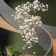 An elegant spiral chandelier featuring white glass petals and gold-toned branches, suspended in a modern stairwell with curved walls. The chandelier has an organic, tree-like structure that twists downward in a graceful helix. Numerous white glass elements, resembling stylized flowers or leaves, are attached to the golden metallic framework, creating a cascading effect. The fixture's warm glow contrasts beautifully with the dark green wall behind it. A portion of a curved, modern staircase with light-colored walls is visible, mirroring the chandelier's spiral form. At the bottom of the image, glimpses of plants and recessed floor lighting add to the luxurious ambiance. The chandelier's intricate design harmonizes with the curved architecture, creating a stunning focal point in this contemporary interior space.