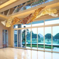 A stunning custom lighting hotel chandelier dominates a luxurious lobby with floor-to-ceiling windows overlooking a tropical landscape. The chandelier features a flowing, wave-like design composed of thousands of glass elements in various golden hues, ranging from deep amber to light gold and clear. The undulating form stretches across the ceiling, creating a sense of movement and opulence. Above the main chandelier, a band of blue and white lighting adds a sky-like effect. The warm, golden glow of the chandelier contrasts beautifully with the cool, natural scenery outside, which includes palm trees, a manicured lawn, and a glimpse of a pool. The polished floor reflects the chandelier's brilliance, enhancing the overall luxurious atmosphere of the space.