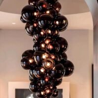 A dramatic custom lighting installation featuring a large hotel chandelier composed of multiple black glass spheres. This big chandelier showcases an array of glossy, opaque orbs clustered together in a cascading formation. The custom lighting piece hangs from the ceiling, creating a bold, sculptural presence against a neutral background. Some spheres emit a warm glow, adding depth and intrigue to the design. The chandelier's unique composition of dark, reflective surfaces and subtle illumination makes it a captivating focal point, perfect for adding contemporary elegance to luxurious spaces.