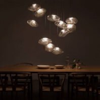 A striking custom lighting installation hangs above a long wooden dining table in a dimly lit room. The lighting features multiple organically shaped glass elements suspended at varying heights, resembling illuminated pebbles or water droplets. Each glass piece emits a soft, warm glow, creating pools of light that stand out dramatically against the dark background. The translucent nature of the glass allows the light to diffuse gently, giving each piece a ethereal, cloud-like quality. The arrangement of the lights is asymmetrical and dynamic, adding visual interest and depth to the space. Below, the wooden dining table is set with simple, elegant dishware and surrounded by minimalist chairs, emphasizing the modern and sophisticated aesthetic. This innovative lighting design transforms the dining area into an intimate, atmospheric space, perfect for a high-end restaurant or a luxurious home dining room seeking to create a memorable ambiance.