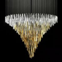 A stunning large chandelier showcasing innovative custom lighting design. This chandelier features an inverted pyramid shape composed of hundreds of elongated crystal-like elements. The top layer consists of clear or white crystals, gradually transitioning to golden hues towards the bottom, creating a mesmerizing ombre effect. The crystals appear to be suspended by thin wires, giving the illusion of a floating, shimmering mass. Set against a dark background, the chandelier's brilliance is accentuated, making it a true statement piece. This luxurious custom lighting installation is perfect for high-end hotels or grand spaces seeking a breathtaking, contemporary centerpiece that combines the opulence of traditional crystal chandeliers with modern design sensibilities.