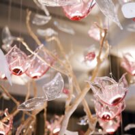 A close-up of a large custom chandelier, perfect for luxury hotel spaces. This big chandelier features an array of delicate glass elements resembling flower petals or leaves. The glass pieces are suspended on thin wires, creating a floating effect. Some elements are clear while others have a soft pink hue, adding depth and warmth. Interspersed among the glass petals are golden branch-like structures, enhancing the organic, nature-inspired design. The interplay of light on the various glass shapes creates a mesmerizing, ethereal glow. This bespoke lighting installation showcases the artistry in custom chandelier design for upscale interiors.