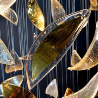 A detailed view of a large custom chandelier, perfect for upscale hotel lobbies. This big chandelier features an array of suspended glass elements resembling leaves or petals. The focal point is a large, amber-tinted glass leaf with a clear edge, creating a striking contrast. Surrounding it are smaller glass pieces in various shapes and textures, some clear with bubble-like inclusions, others with golden hues. Thin wires delicately suspend each element, adding to the chandelier's ethereal quality. This bespoke lighting fixture showcases the artistry and innovation in custom lighting design for luxury spaces.