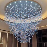 A breathtaking large chandelier showcasing innovative custom lighting design for upscale hotels. This big chandelier features a circular base with numerous LED lights, from which thousands of crystal droplets in clear and light blue hues are suspended. The hotel chandelier creates a stunning waterfall effect, with crystals densely clustered at the bottom and gradually dispersing towards the top. This exquisite custom lighting installation transforms the space into a mesmerizing display of cascading water droplets, perfect for creating a serene and luxurious atmosphere in high-end hospitality environments.