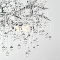 A detailed view of a modern chandelier featuring an intricate design of glass elements. The structure consists of intertwining branches or stems made of clear glass, resembling a crystalline tree or abstract sculpture. Hanging from these branches are numerous thin glass rods of varying lengths, each terminating in a small spherical glass droplet. These droplets create a cascading effect, reminiscent of raindrops or icicles. The glass elements are transparent and reflective, catching and refracting light. In the background, a warm glow from the chandelier's light source is visible, illuminating the glass pieces and creating a sparkling effect. The overall design is organic yet contemporary, blending natural inspiration with sleek, modern aesthetics. This chandelier would serve as a striking focal point in a modern or luxury interior space.