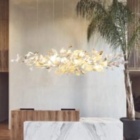 A long, horizontal chandelier with illuminated flower-like shapes hangs above a marble-topped counter. The fixture resembles a glowing branch with delicate petals and leaves. A potted palm tree stands to the right, adding a tropical touch. The room features neutral tones with wood accents, creating a harmonious blend of natural and modern elements. The chandelier serves as a striking focal point in this elegant, nature-inspired interior.