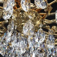 Close-up of a luxurious chandelier featuring gold-plated floral designs and numerous crystal droplets. Intricate gold patterns resemble blooming flowers, while faceted crystal pendants hang below, refracting light in rainbow hues. The combination creates a dazzling, opulent effect.