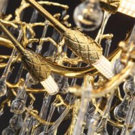 A detailed close-up of a luxurious chandelier featuring intricate gold-plated leaf designs and crystal elements. The image showcases two prominent gold leaves with textured surfaces and veining, attached to curved golden branches. These leaves appear to be light fixtures, with small LED or bulb bases visible. Surrounding the gold elements are numerous crystal droplets and rods of varying lengths, creating a cascading effect. The crystal pieces reflect and refract light, adding sparkle and brilliance to the overall design. The combination of warm gold tones and clear crystals creates a rich, opulent aesthetic. This chandelier exemplifies a fusion of nature-inspired motifs with high-end materials, suitable for elegant and luxurious interior spaces.