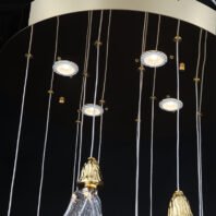 Close-up of a contemporary branch chandelier featuring a mix of lighting elements. Thin silver wires suspend glass leaf-shaped pendants with gold bell-shaped caps, partially visible at the bottom. Interspersed among these are modern LED spotlights with clear glass surrounds. The chandelier's design combines organic, nature-inspired forms with sleek, modern lighting technology. Gold accents on the wires and caps add a touch of elegance. This branch chandelier showcases a unique blend of natural and contemporary aesthetics, suitable for modern interiors seeking a balance of artistry and functionality.