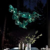 Mesmerizing branch chandelier featuring illuminated teal glass leaves suspended over a dining table. The chandelier's organic design mimics a floating canopy of glowing foliage, each leaf-shaped element lit from within. Set against a dark night sky visible through large windows, the chandelier creates a magical ambiance. Below, a stylish table setting with floral arrangements and decorative vases complements the nature-inspired lighting, showcasing the chandelier's ability to transform an evening dining space.