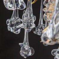 A close-up view of exquisite hand-blown glass pendants from a chandelier, showcased against a dark background. The pendants feature intricate, organic shapes reminiscent of abstract flowers or marine life. Each glass element is uniquely formed, with swirling, flowing lines and bulbous protrusions that create a sense of movement and fluidity. The glass is clear and translucent, allowing light to pass through and create subtle refractions and reflections. The stems of the pendants are long and slender, transitioning into more elaborate forms at their ends. Some pendants appear to have multiple layers or folds in the glass, adding depth and complexity to their design. In the background, glimpses of gold-toned metal can be seen, likely part of the chandelier's structure. The image also captures a small portion of an LED light, visible as a series of small yellow dots, hinting at how the chandelier would illuminate these delicate glass forms. This chandelier combines artisanal glasswork with modern lighting design, resulting in a piece that is both a functional light fixture and a stunning sculptural element.