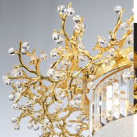 A detailed view of an exquisite chandelier featuring intricate gold-plated branches adorned with crystal elements. The image showcases a complex network of golden branches that twist and intertwine in organic, tree-like formations. These branches are meticulously crafted with a smooth, polished finish that gleams brilliantly. Attached to the golden branches are numerous faceted crystal orbs of varying sizes, resembling sparkling dewdrops or blossoms. These crystals are precisely cut, catching and refracting light to create a dazzling display. The crystals are strategically placed at the ends of branches and at junction points, enhancing the natural, floral-inspired design. In the lower right corner of the image, a portion of the chandelier's main structure is visible, featuring a textured gold band and a series of rectangular crystal prisms hanging vertically. These prisms add an additional layer of light refraction and sparkle to the overall design. The combination of the warm, lustrous gold with the clear, sparkling crystals creates a luxurious and elegant aesthetic. This chandelier beautifully merges nature-inspired design with opulent materials, resulting in a piece that is both a functional light fixture and a stunning work of art, perfect for high-end interior spaces.