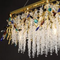 An opulent rectangular chandelier featuring a stunning combination of gold, clear crystals, and colorful gemstone-like accents. The fixture is suspended from a polished gold rectangular frame. From this frame, intricate gold branches extend downward, adorned with multi-colored crystal flowers in hues of blue, turquoise, amber, and clear. These vibrant crystal accents provide pops of color against the predominantly gold and clear crystal design. The most striking feature is the abundant cascade of clear crystal strands hanging from the gold frame, creating a shimmering curtain effect. These crystal strands are arranged in varying lengths, giving the chandelier a waterfall-like appearance. The clear crystals catch and refract light, producing a dazzling sparkle. The chandelier is illuminated from within, causing the entire piece to glow warmly against the dark background. This lighting fixture masterfully combines the warmth of gold with the cool brilliance of crystals, resulting in a piece that is both a functional light source and a luxurious work of art, capable of making a dramatic statement in any high-end interior space.