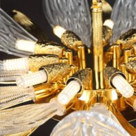 A close-up view of an opulent branch chandelier featuring a blend of gold and crystal elements. The central structure is made of polished gold-toned metal, with intricate detailing resembling a floral stem. Multiple LED light bulbs protrude from ornate gold casings, each designed to look like a blooming flower bud. These LED lights are visible, emitting a warm glow. Surrounding the gold structure are large, petal-like crystal elements with intricate textures reminiscent of feathers or leaf veins. These transparent crystal petals catch and refract light, creating a sparkling effect. The combination of the warm gold tones, the cool crystal elements, and the illuminated LED bulbs creates a stunning visual contrast. This branch chandelier masterfully merges modern lighting technology with nature-inspired luxury design, showcasing the intricate craftsmanship in every detail. The image captures the chandelier's ability to serve as both a functional light source and a piece of sculptural art in high-end interiors.