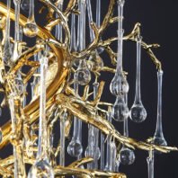 A close-up view of an exquisite branch chandelier showcasing intricate gold-plated metalwork intertwined with crystal elements. The chandelier's structure features elaborately designed golden branches that twist and turn organically, mimicking the natural growth patterns of trees. These polished gold-toned branches create a striking contrast against a dark background. Suspended from the golden framework are numerous clear crystal pendants of varying lengths, each consisting of a slender stem ending in a bulbous teardrop shape. These crystal elements resemble dewdrops or raindrops, adding a ethereal quality to the design. The interplay between the warm, reflective gold surfaces and the cool, transparent crystal droplets creates a mesmerizing visual effect. This branch chandelier masterfully blends luxurious materials with nature-inspired forms, resulting in a stunning piece that bridges the gap between organic beauty and high-end interior design.