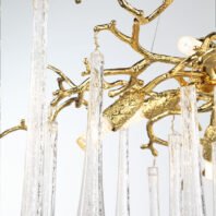 A close-up view of an exquisite branch chandelier featuring golden, intricately designed branches reminiscent of a tree. The chandelier's structure is crafted from polished gold-toned metal, with realistic textures mimicking tree bark and twigs. Suspended from these golden branches are multiple long, slender crystal pendants that resemble icicles or raindrops. These clear crystal droplets catch and refract light, creating a sparkling effect against the warm gold tones. This branch chandelier seamlessly blends organic, nature-inspired forms with luxurious materials, offering a stunning focal point for sophisticated interiors. The image captures the chandelier's delicate balance between the solid, gleaming gold branches and the fragile, transparent crystal elements.