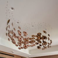 An innovative custom lighting installation featuring a swirling arrangement of copper-colored discs suspended from a white ceiling. The oval-shaped elements vary in size and are hung at different heights, creating a dynamic, wave-like pattern that flows across the ceiling. The highly polished, reflective surfaces of the discs capture and reflect light, producing a warm, metallic glow that ranges from rose gold to deep bronze hues. Thin, barely visible wires support each disc, giving the illusion of floating metallic bubbles. This unique lighting design transforms the space into an artistic display of light and movement, perfect for upscale hotel lobbies, high-end restaurants, or luxurious event venues. The installation showcases how bespoke lighting can serve as both functional illumination and a striking architectural feature in sophisticated interiors.