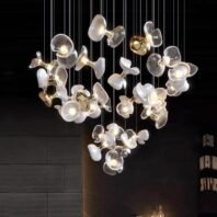 A stunning large chandelier showcasing innovative custom lighting design for upscale hotels. This big chandelier features a cluster of translucent white glass elements shaped like abstract flower petals or shells. Each petal-like piece is illuminated from within, creating a soft, pearlescent glow against a dark background. The hotel chandelier's unique composition resembles a floating cloud of delicate blossoms, suspended at varying heights on thin wires. Gold accents connect some glass pieces, adding a touch of warmth and luxury. This exquisite custom lighting installation transforms the space into an enchanting, nature-inspired display, perfect for creating a sophisticated and modern atmosphere in high-end hospitality environments.